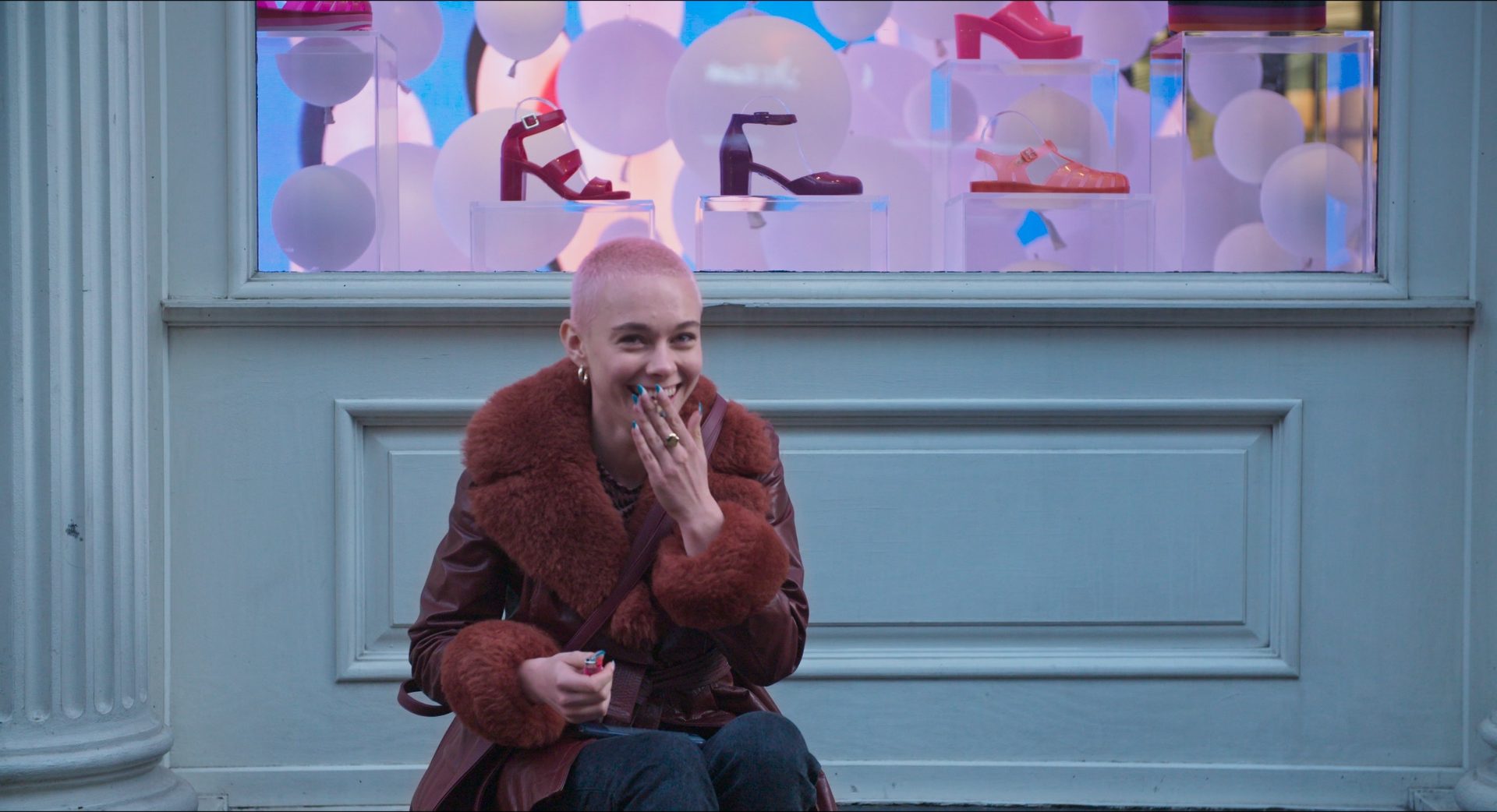 A young woman with a cropped pink buzz cut and a rust fur-trimmed coat laughs behind her hand, sitting in front of a pastel shop window.