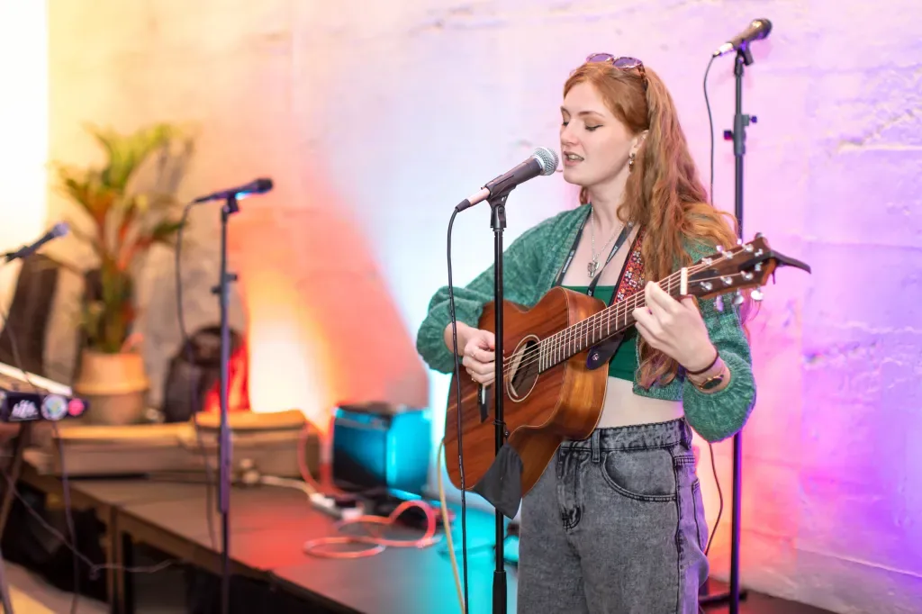 Lottie Hartnack sings and plays acoustic guitar at a microphone, lit in soft pink and amber.