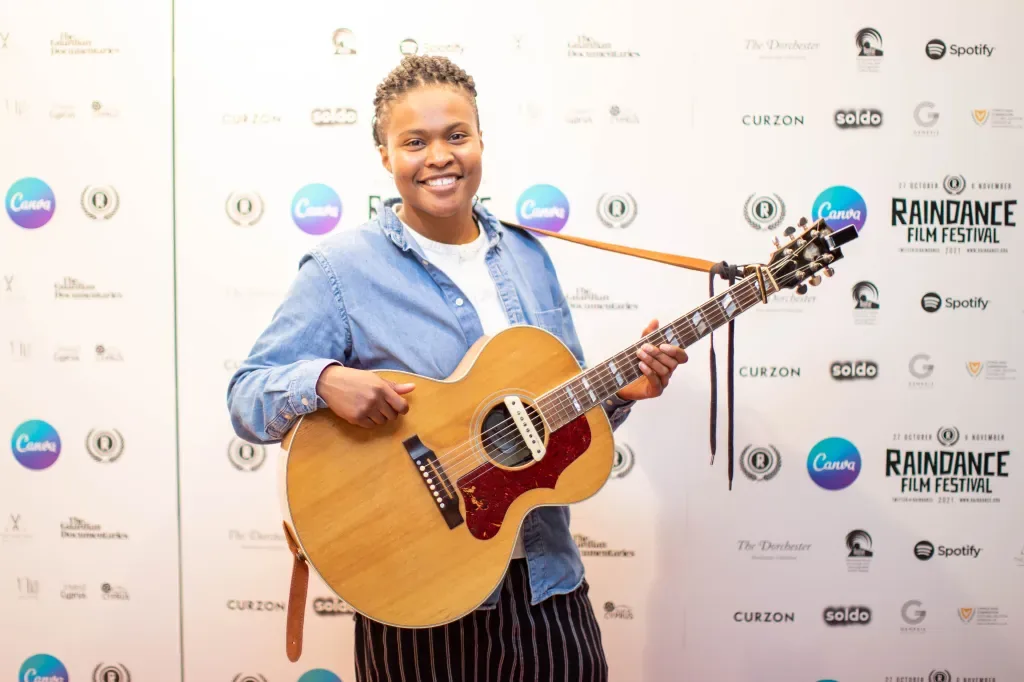 Sherika Sherard smiles holding an acoustic guitar in front of the Raindance Film Festival press wall.